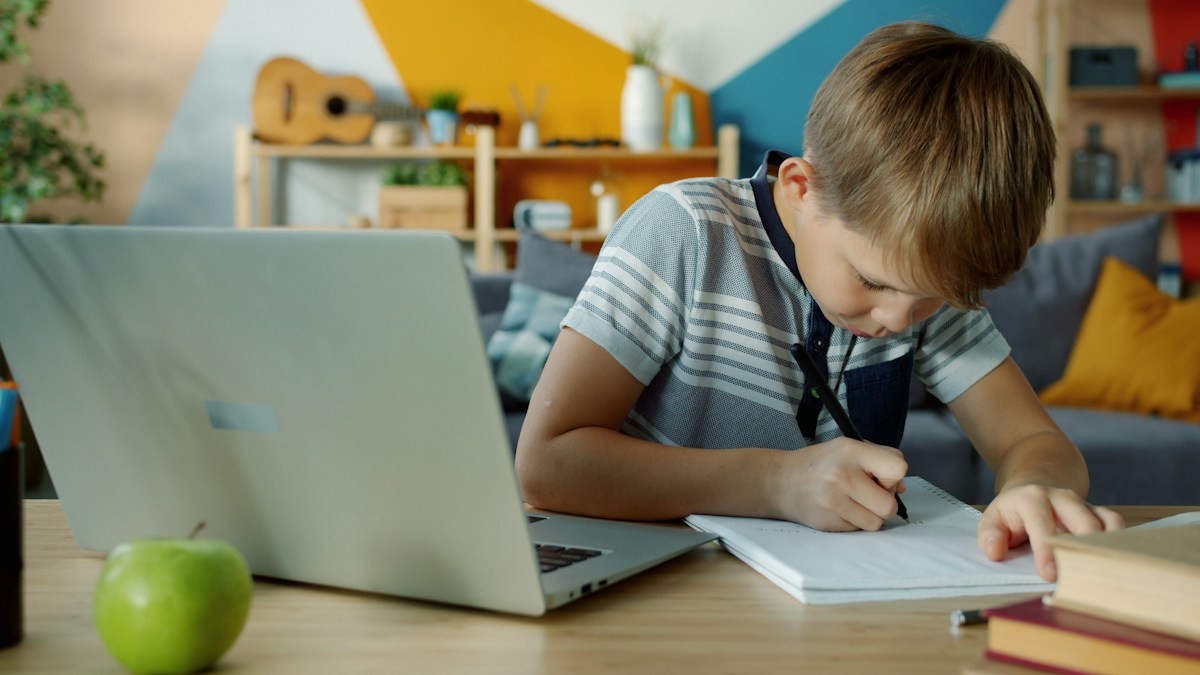 A school student learning on a laptop and writing in his notebook